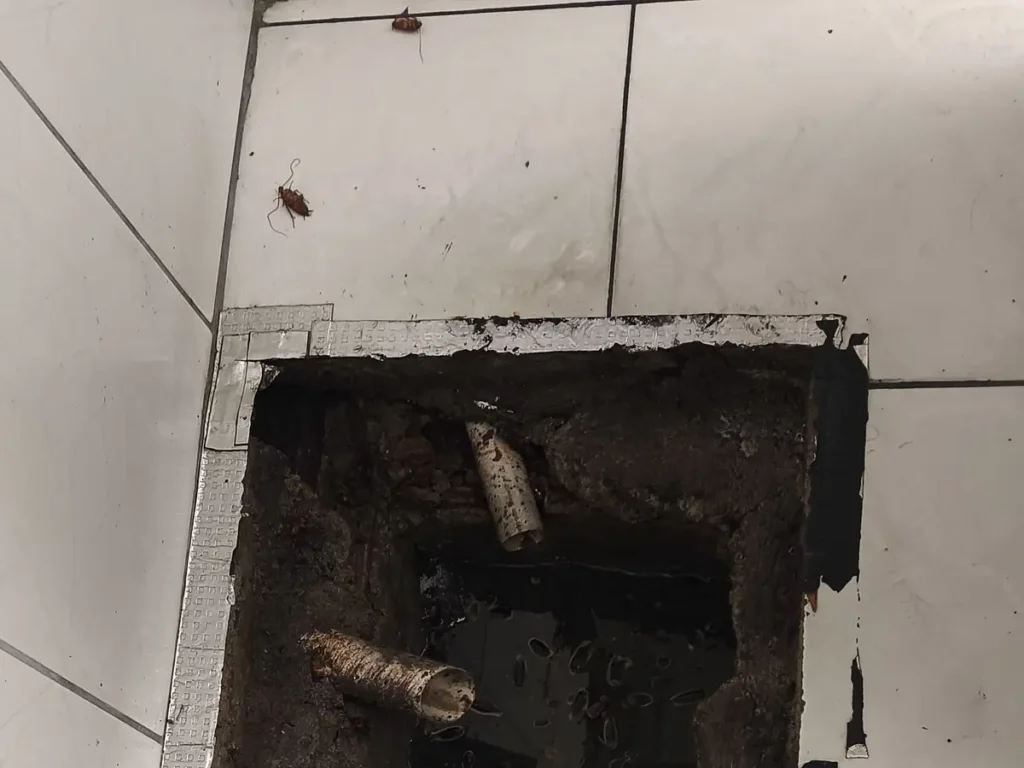 Caixa de inspeção de esgoto aberta no piso, com os tubos de entrada e saída à vista